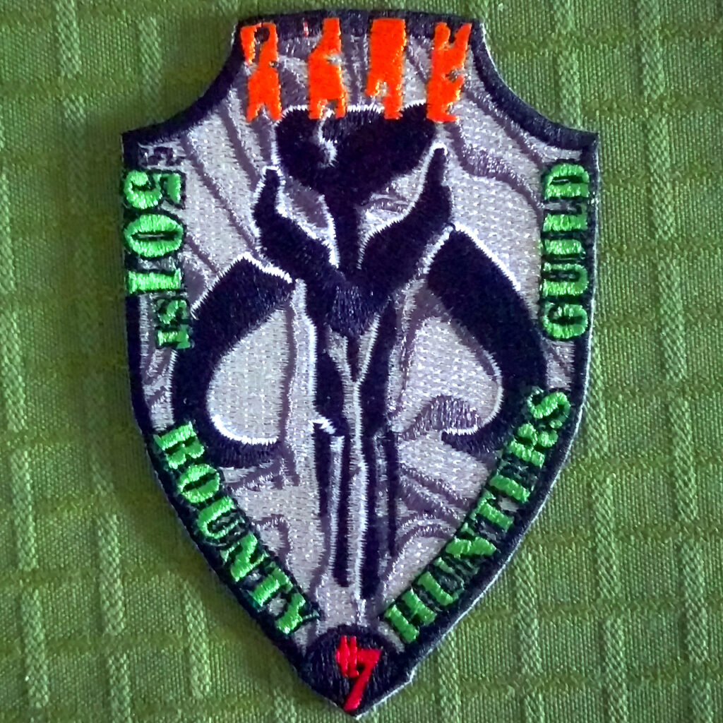 An embroidered patch shaped like a shield. Black borders all around the shield. On top four orange lines representing "kill stripes". On the bottom a red aurabesh symbol representing credits/money. In the middle a large "mythosaur" skull symbol. Grey, reflective background. Text in green down on the sides: "501st Bounty Hunters Guild"