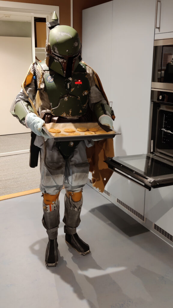Boba Fett is in a kitchen. He has oven mittens on, and has pulled a baking tray out of the oven. The tray is filled with golden brown pastries shaped like spirals.