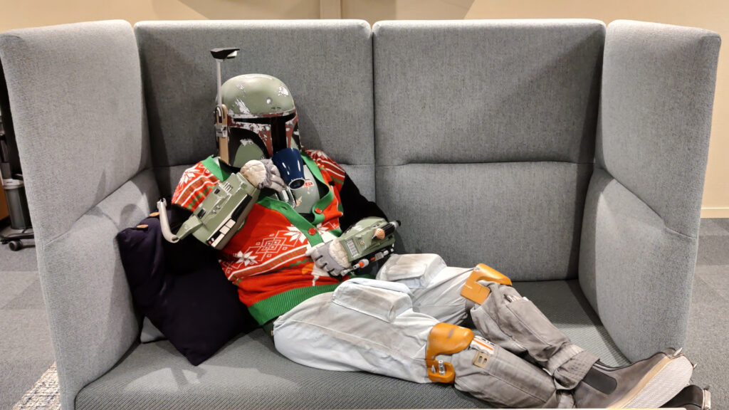 Boba Fett relaxes on the couch. He has a festive sweater on, and drinks hot chocolate out of a mug.