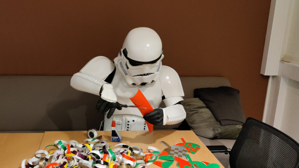A stormtrooper sits at a table, making christmas decorations out of glossy paper. He holds a piece of paper in one hand and scissors in the other, looking very concentrated.