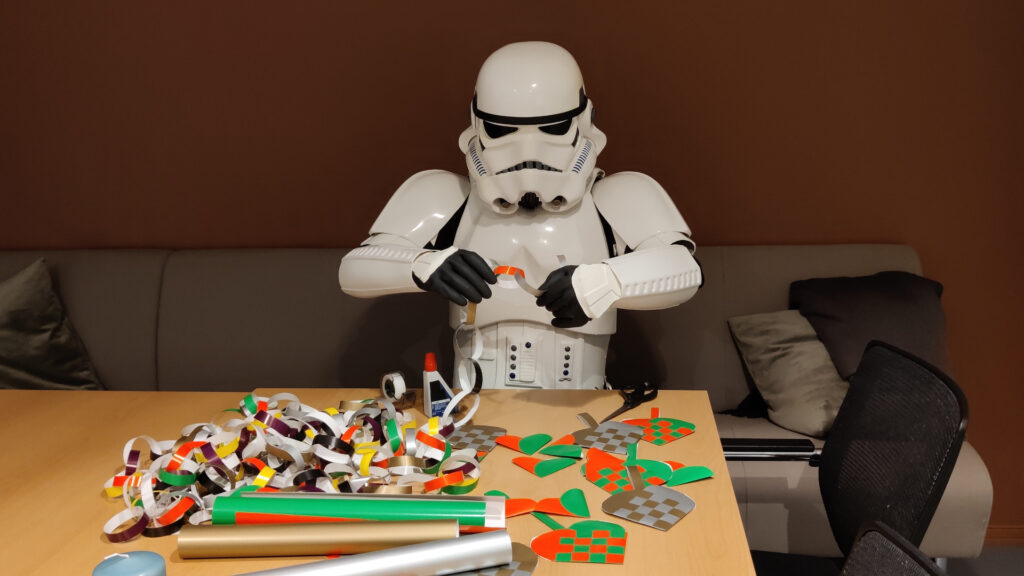 A stormtrooper sits at a table making christmas decoration out of glossy paper. He is threading a paper onto a long paper chain.