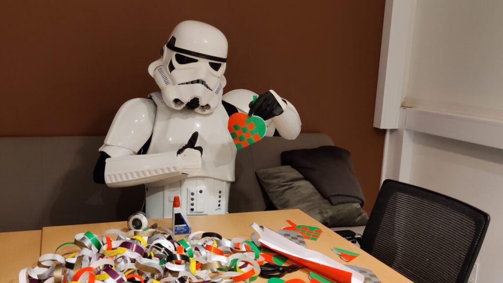 A stormtrooper sits at a table, making christmas decorations out of glossy paper. He holds up a weaved christmas heart with one hand and gives a thumbs up with the other.