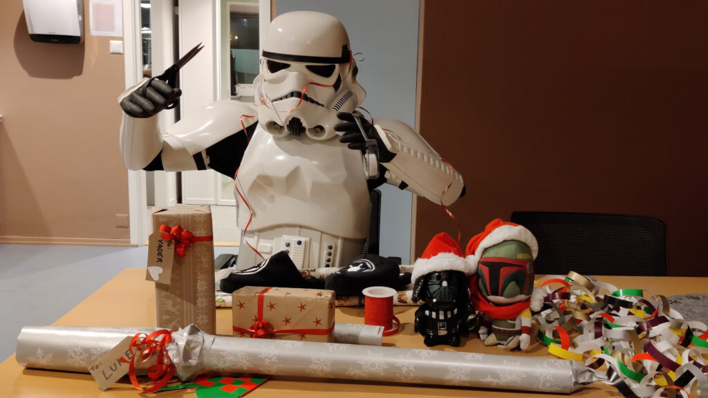 A stormtrooper sits at a table, wrapping christmas gifts. A few gifts has already been wrapped. He is now wrapping imperial slippers, but struggles with the tape and the gift ribbon.