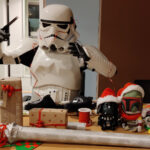 A stormtrooper sits at a table, wrapping christmas gifts. A few gifts has already been wrapped. He is now wrapping imperial slippers, but struggles with the tape and the gift ribbon.