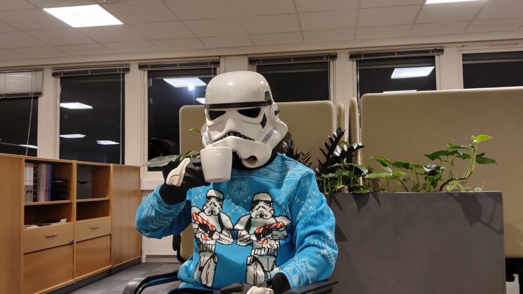 A stormtrooper wearing a festive sweater, sitting in an office drinking from a coffee mug.