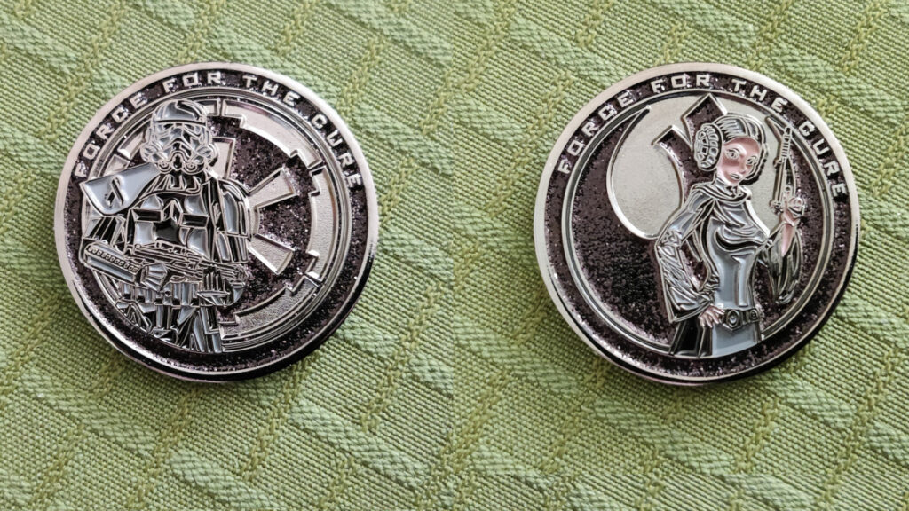 A black coin. We see both faces side by side. One side features the Imperial cog symbol and an Imperial Stormtrooper. The other side features the Rebel Alliance Starbird insignia and Princess Leia. Both sides says "Force for the cure" on top.