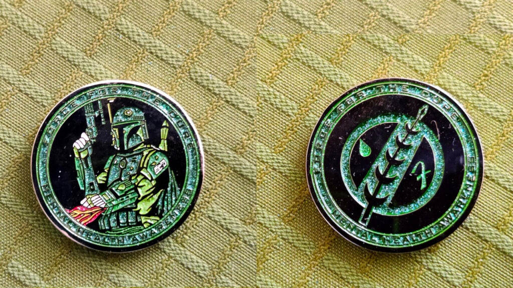 A sparkly green and black coin. We see both faces side by side. One side features Boba Fett. The other side features his Jaster feather insignia. Both sides reads "Force for the cure" and "Mental Health Awaresness".