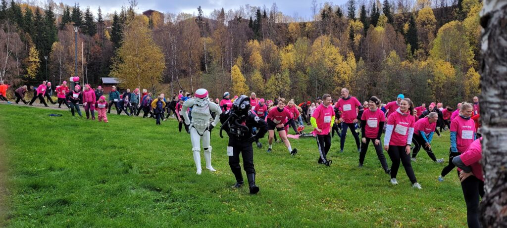 A large group of people in a park are doing a warm-up. Most are dressed in pink. All are wearing starting numbers for the Pink Ribbon Charity Run. A stormtrooper and a TIE pilot is among the crowd.