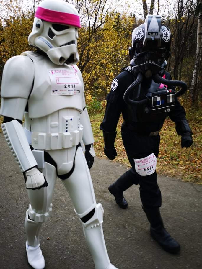 A stormtrooper and a TIE pilot on a pathway. Both are wearing starting numbers for the Pink Ribbon Charity Run. The Stormtrooper are also wearing a pink headband. 