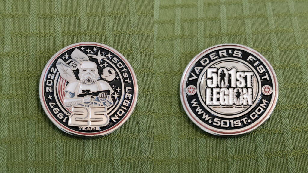 A round metal coin. Front: A red border. In the middle we see a stormtrooper standing guard with their E11 Blaster Rifle. Behind them we see a TIE Fighter with red engine streaks, the Death Star and several stars in space. The text going around the Stormtrooper in a circle reads: "1997-2022 - 501st Legion - 25 Years". The outlines of the drawing is in gold. Back: A red border. The 501st Letter Logo with two imperial cogs on each side, with the text "Vader's Fist" and "www.501st.com" along the border.