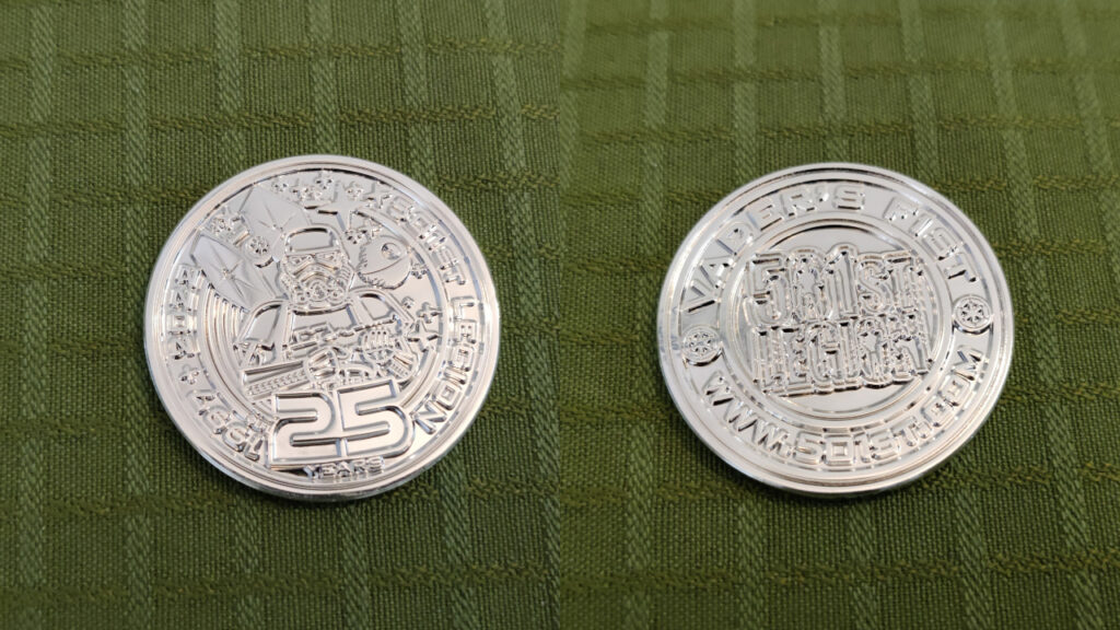 A round, silver plated, metal coin. Front: In the middle we see a stormtrooper standing guard with their E11 Blaster Rifle. Behind them we see a TIE Fighter with engine streaks, the Death Star and several stars in space. The text going around the Stormtrooper in a circle reads: "1997-2022 - 501st Legion - 25 Years". Back: The 501st Letter Logo with two imperial cogs on each side, with the text "Vader's Fist" and "www.501st.com" along the border.