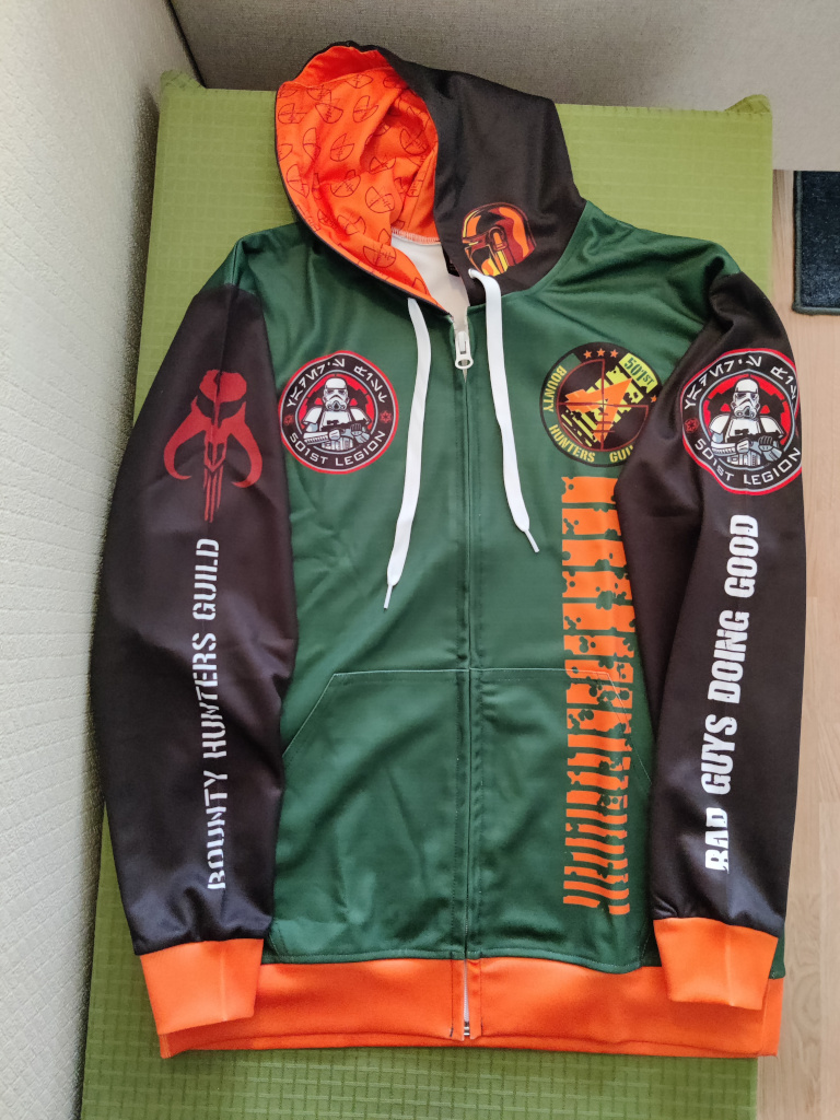 The jacket flat on a table. We see that it is a green jacket with black sleeves and hoodie, white cords and orange trim. One arm has the Mythosaur logo and the text "Bounty Hunters Guild". The other arm has the round 501st Logo and the text "Bad Guys Doing Good". The right side of the chest has the round 501st Logo. The left side of the chest has the round Bounty Hunters Guild Logo. Down the left side is the orange kill stripes from Boba Fett's helmet.