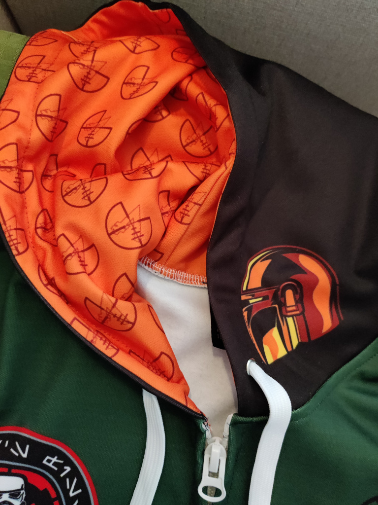 Close-up of the black hoodie. It has an orange mandalorian helmet on the outside. The inside is lined in orange fabric with the Bounty Hunters Guild Logo crosshairs as a pattern.