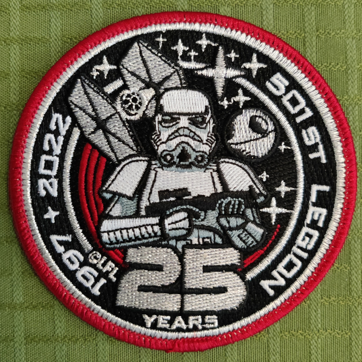 A round embroidered patch with a red and white border. In the middle we see a stormtrooper standing guard with their E11 Blaster Rifle. Behind them we see a TIE Fighter with red engine streaks, the Death Star and several stars in space. The text going around the Stormtrooper in a circle reads: "1997-2022 - 501st Legion - 25 Years".