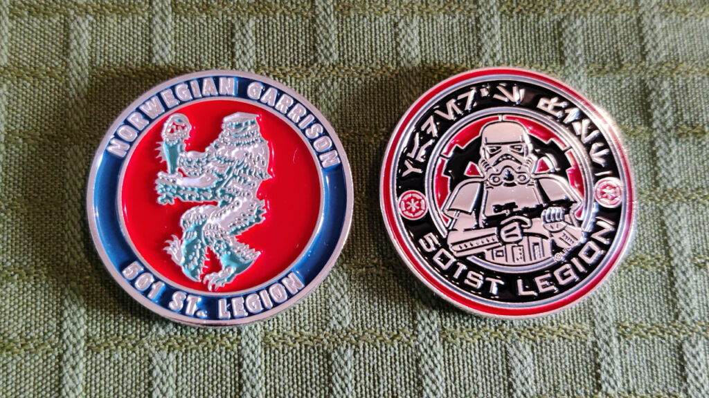 A silver metal coin, front and back. The border of the coin is blue with silver letters that reads "501st Legion" and "Norwegian Garrison". The face of the coin is a stylized figure against a red background. It is a wampa, the ice monster on planet Hoth from the movie Star Wars: Episode V – The Empire Strikes Back. The wampa is walking to the left while carrying a tauntaun leg. The style and the pose bears a resemblance to the Coat of Arms of Norway.  The back of the coin is the round 501st Legion logo, with a red and black border that reads "501st Legion" in latin letters and "Vader's Fist" in aurebesh letters. The face is a stormtrooper.