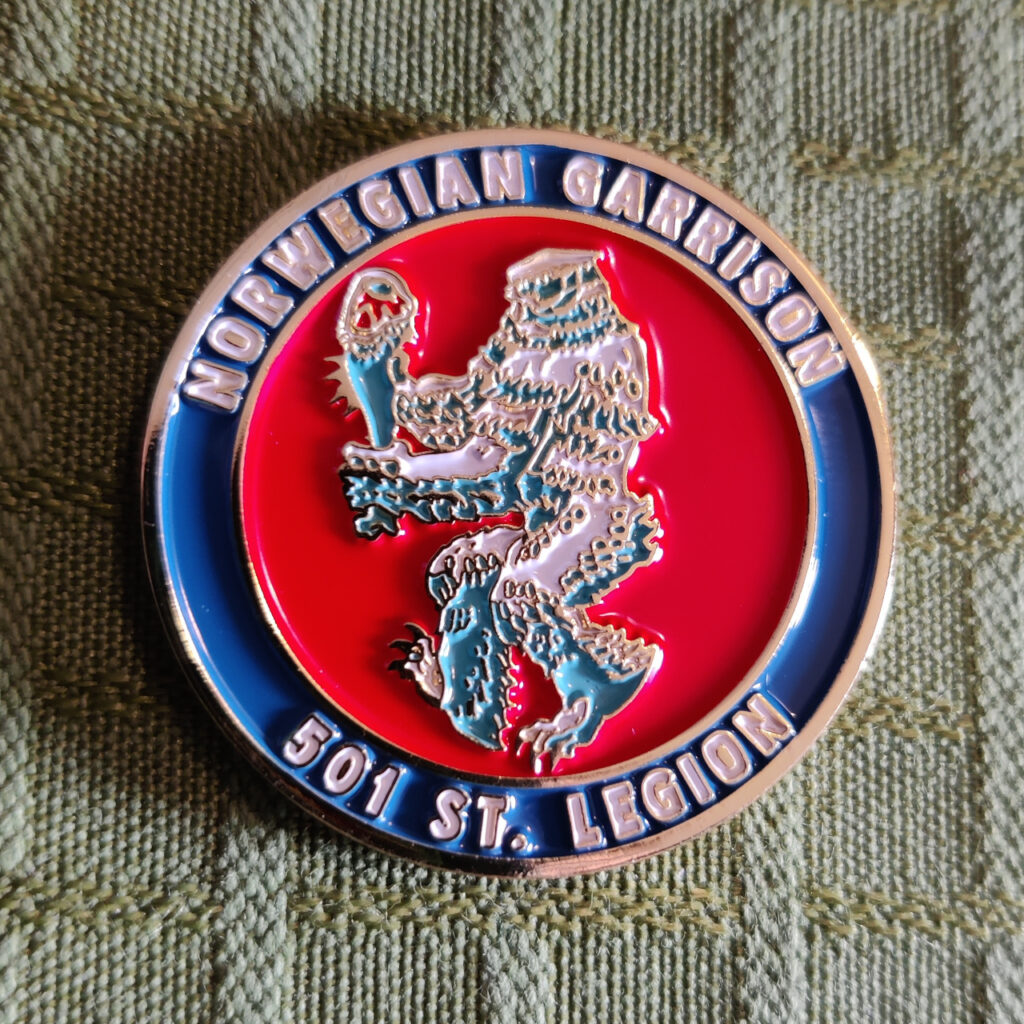 A gilded metal coin. The border of the coin is blue with gold letters that reads "501st Legion" and "Norwegian Garrison". The face of the coin is a stylized figure against a red background. It is a wampa, the ice monster on planet Hoth from the movie Star Wars: Episode V – The Empire Strikes Back. The wampa is walking to the left while carrying a tauntaun leg. The style and the pose bears a resemblance to the Coat of Arms of Norway. All highlights is gold.