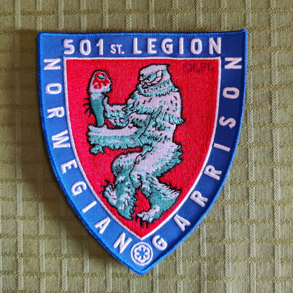 A embroidered patch in the shape of a shield. The border of the shield is blue with white letters that reads "501st Legion" and "Norwegian Garrison". The face of the shield is a stylized figure against a red background. It is a wampa, the ice monster on planet Hoth from the movie Star Wars: Episode V – The Empire Strikes Back. The wampa is walking to the left while carrying a tauntaun leg. The style and the pose bears a resemblance to the Coat of Arms of Norway.