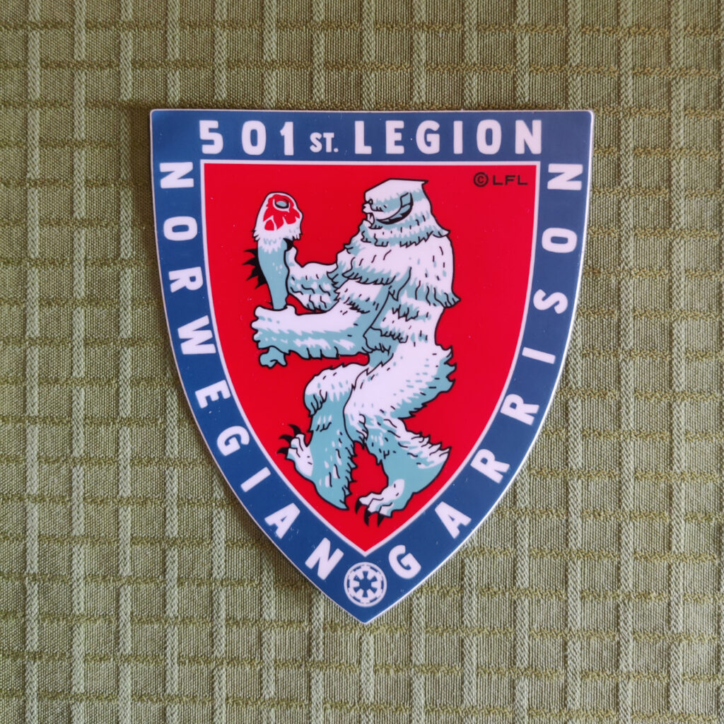 A sticker in the shape of a shield. The border of the shield is blue with white letters that reads "501st Legion" and "Norwegian Garrison". The face of the shield is a stylized figure against a red background. It is a wampa, the ice monster on planet Hoth from the movie Star Wars: Episode V – The Empire Strikes Back. The wampa is walking to the left while carrying a tauntaun leg. The style and the pose bears a resemblance to the Coat of Arms of Norway.