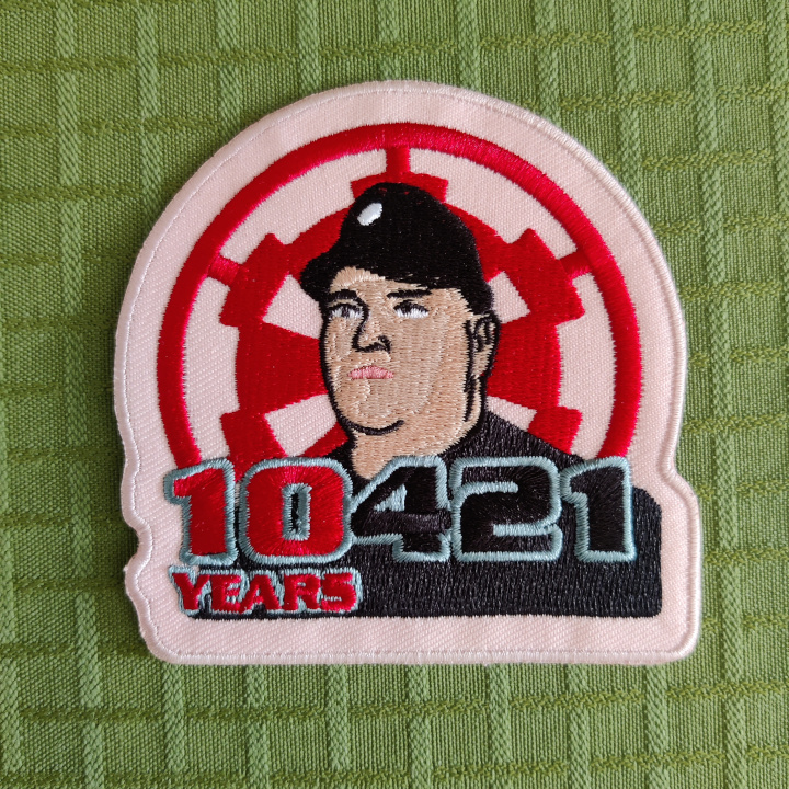 An embroidered patch. 

The graphics is a realistic drawing of a man in an imperial officer uniform, looking to the left. 

The background is the imperial cog. 

The letters on the bottom reads "10421 years", with "10" and "years in the same color.