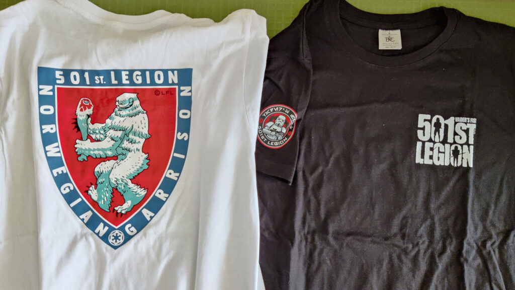 Two t-shirts on a table. 
The shirt on the left is white, and has a large Norwegian Garrison logo. 
The shirt on the right is black,  has the round 501st Legion logo on the sleeve and the 501st Legion letter logo on the chest.