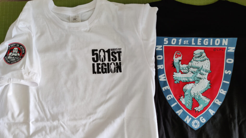Two t-shirts on a table. 
The shirt on the left is white, has the round 501st Legion logo on the sleeve and the 501st Legion letter logo on the chest. 
The shirt on the right is black, and has a large Norwegian Garrison logo. 