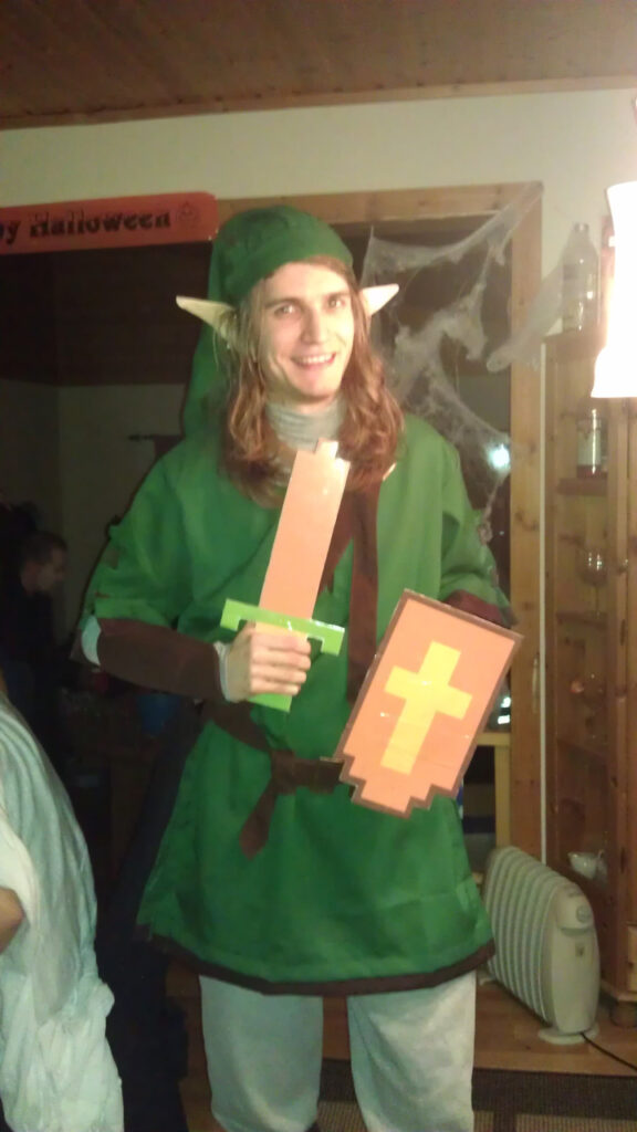 I'm dressed as Link from the first Zelda game. The costume is a green tunic with brown vambraces and a belt, grey pants, grey undershirt, and a long green hat. I am holding a 8 bit sword and an 8 bit shield with a cross, just like in the game. I have long hair and pointy ears. The hear is mine, the ears are costume pieces.