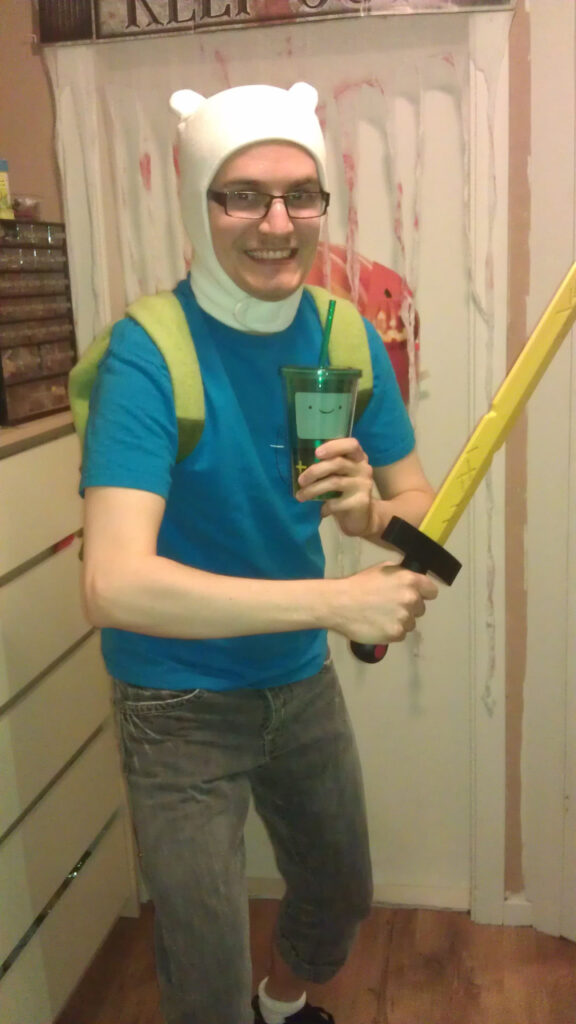 I'm dressed as Finn the Human. It's a blue t-shirt, a pair of knee-high black denim pants, a green backpack and a white hat with "ears". I am holding a green cup with a face (inspired by the robot B-MO from the show) and a yellow sword (from the opening titles of the show). Hidden by the cup is a pocket on the t-shirt where a tiny Jake the Dog is sitting.