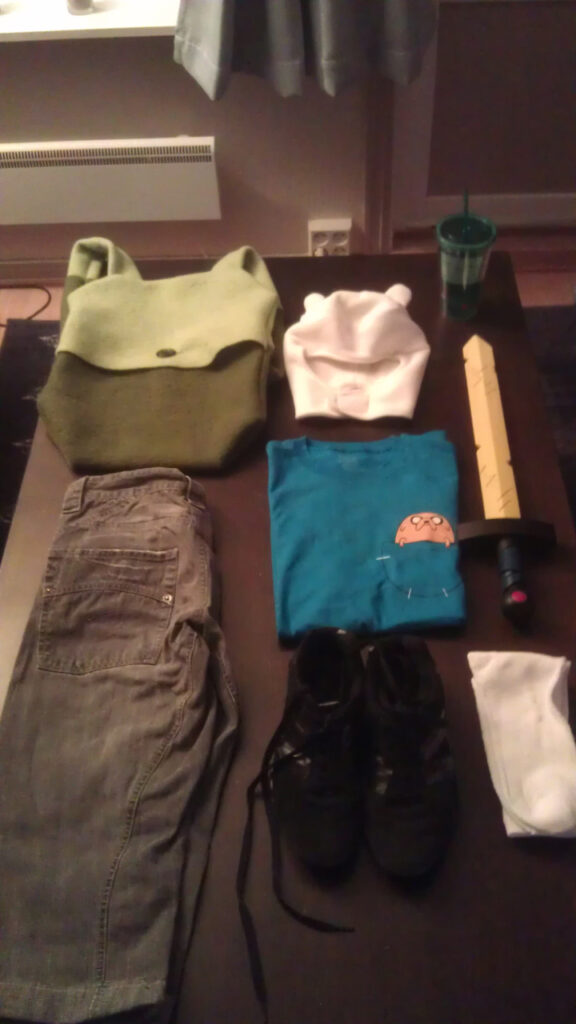 A table with the Finn the Human costume. A green backpack, a hat with "ears", a green cup with a face (inspired by the robot B-MO from the show), a pair of knee-high black denim pants, a blue t-shirt with a pocket where a tiny Jake the Dog is sitting, a yellow sword (from the opening titles of the show), black shoes and white tennis socks.