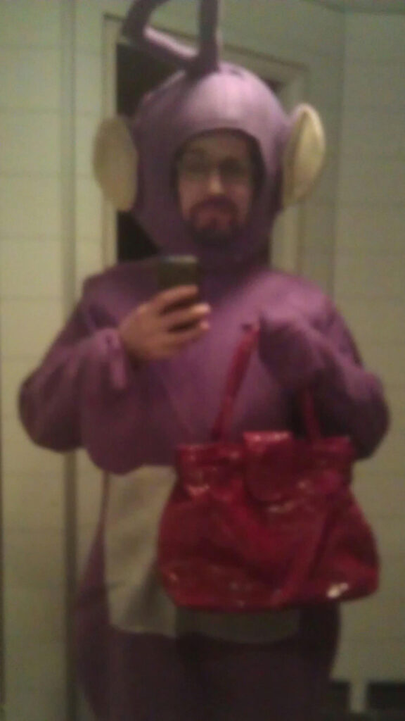 I'm dressed as Tinky Winky from The Teletubbies. It's a full-body purple suit with large ears, a silver tummy and an antenna on the head. I am holding a red purse. I am taking a selfie in a mirror. The image is a bit fuzzy, but we see that I have colored my beard purple to match the suit. (I had to shave the beard afterwards because the temporary color turned out to be permanent.)