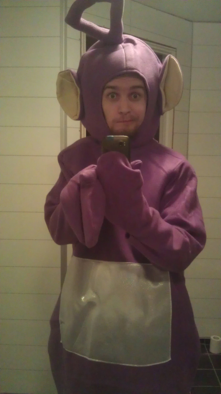 I'm dressed as Tinky Winky from The Teletubbies. It's a full-body purple suit with large ears, a silver tummy and an antenna on the head. I am taking a selfie in a mirror, looking a bit perplexed since it is very difficult to operate a phone with purple mitten hands.