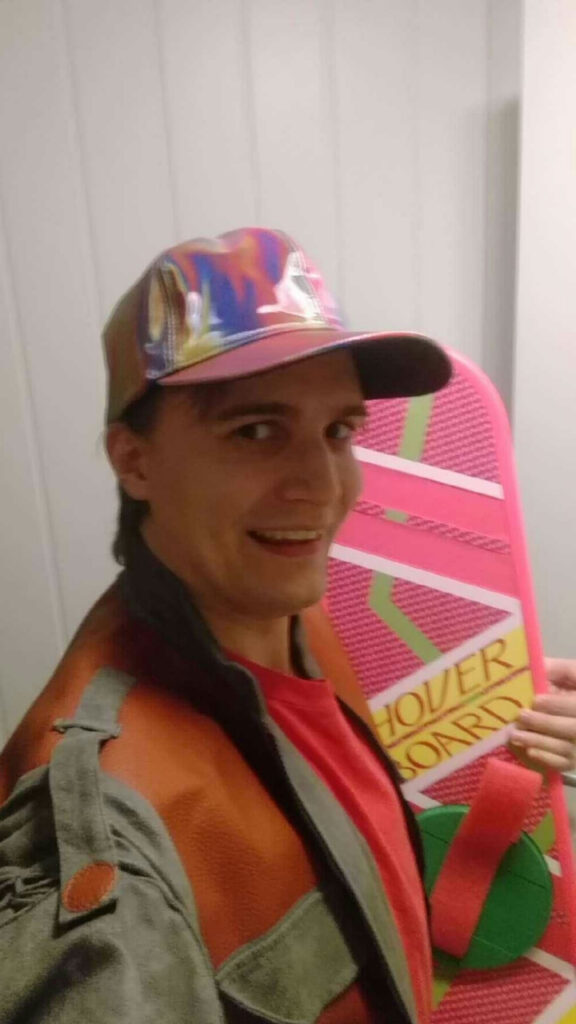 I am dressed as Marty from Back to the Future 2. The costume is a red t-shirt, maroon and grey jacket and a rainbow colored holographic hat.
