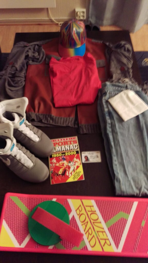 A table with Marty's costume. It's a red t-shirt, a maroon and grey jacket, a rainbow colored holographic hat, a pair of jeans with the pockets on the outside, a pair of futuristic Nike shoes, a 1950–2000 Sports Almanac, Marty's driving license, and a hover board.