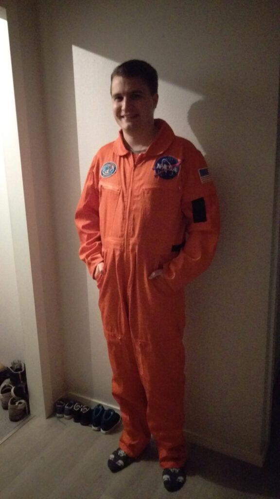 I'm dressed as a NASA employee. It is a orange jumpsuit with NASA patches. My hair is combed. I am standing with my hands in my pockets, giving a wide smile.