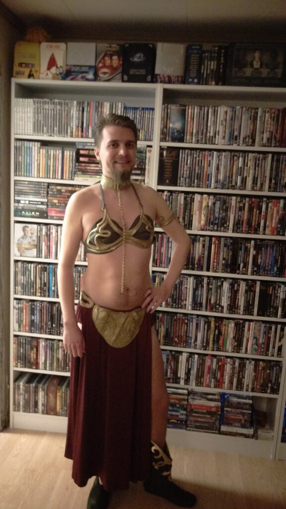 I am dressed as Leia the Huttslayer. The costume is a gold bikini, a maroon skirt with a very high split, boots with gold details and a chain around my neck. Lot's of skin is showing. I am smiling very self-aware and awkwardly.
