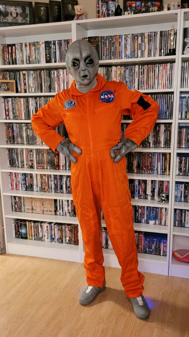 I am dressed as an alien. The costume is an orange jumpsuit with NASA patches and a pair of futuristic Nike shoes. My hands is grey and weird and only has three fingers. I have a full-head mask. The face is grey with all black eyes and a tiny mouth.