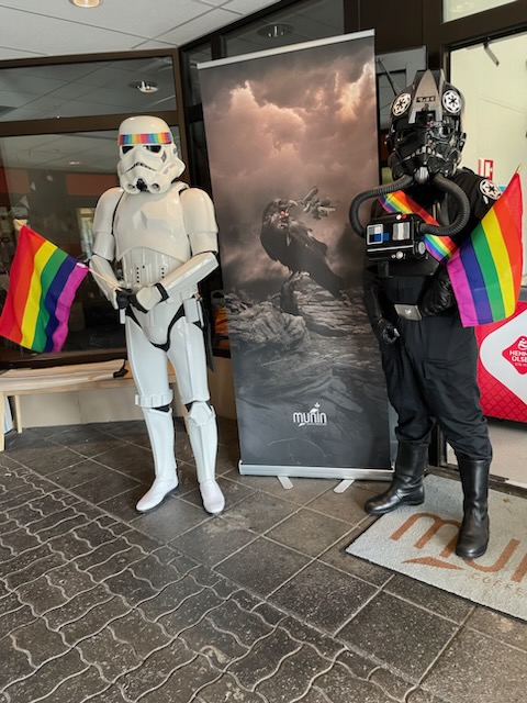A stormtrooper with a rainbow colored head band and a TIE pilot with a rainbow colored sash is standing outside Munin coffeehouse. 