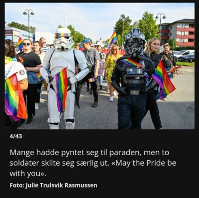A screenshot from the newspaper. We see a huge parade with lots of people. In front of the photographer is a stormtrooper with a rainbow colored head band and a TIE pilot with a rainbow colored sash. Both carries rainbow flags.