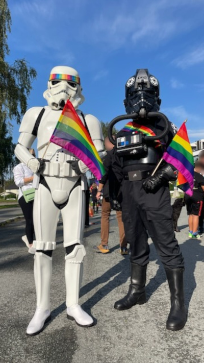 A stormtrooper with a rainbow colored head band and a TIE pilot with a rainbow colored sash is posing for the camera. Both have rainbow flags.