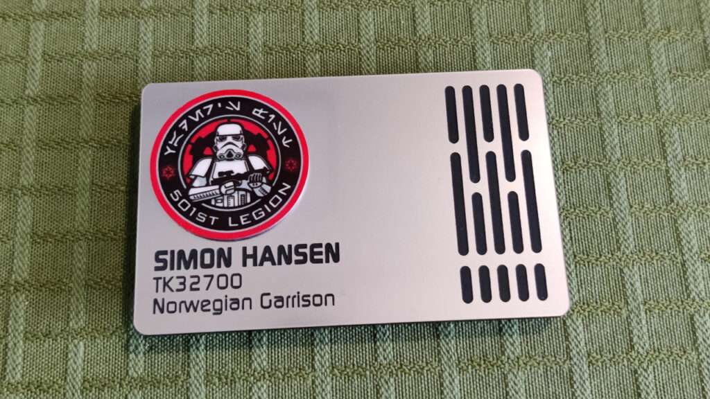 A name badge in faux metal with long, oval cutouts, like the wall panels on the Death Star, backed with black. To the left is a raised 501st Legion logo. 

Text engraved below the logo: 

"Simon Hansen.
TK32700. 
Norwegian Garrison".