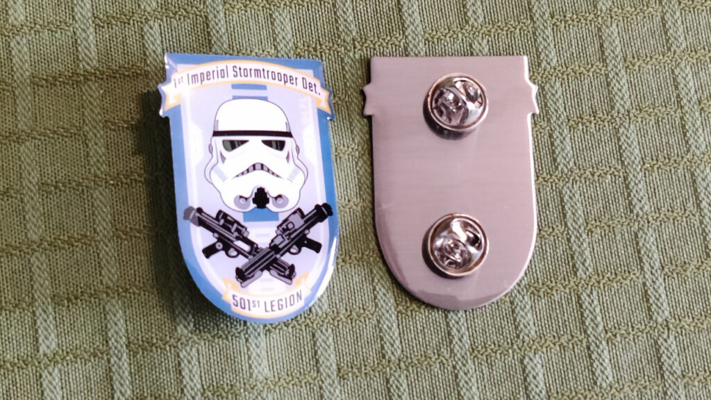 Two lapel pins, back and front. The log is shaped like a shield with the text "1st Imperial Stormtrooper Det." on a banner on top, a stormtrooper helmet in the middle, two E-11 stormtrooper blaster rifles in a cross below the helmet (sharing a similarity to pirate flags), and another smaller banner at the bottom with the text "501st Legion".On the back we see that the pin has two prongs with butterfly clutches.