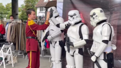 Three stormtroopers are standing in front of a photo wall. Someone wearing a marching band uniform is walking up to them and giving them high-fives.