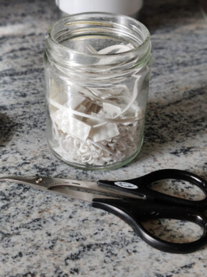 A glass jar filled with small pieces of white ABS plastic. In front of the jar we see a pair of scissors for cutting plastic.