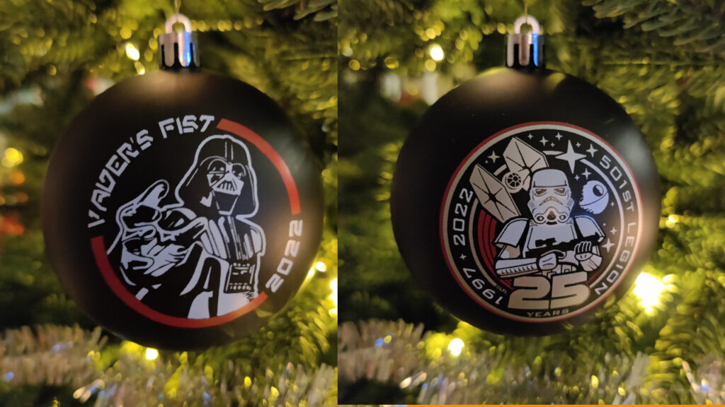 A black ornament hanging in a lit Christmas tree. On the front of the bauble we see a stylized drawing of Darth Vader reaching out to us with the text "Vader's Fist 2022". On the back we see the 501st Legion 25th Anniversary logo. It's a red and white bordered circle. In the middle we see a stormtrooper standing guard with their E11 Blaster Rifle. Behind them we see a TIE Fighter with red engine streaks, the Death Star and several stars in space. The text going around the Stormtrooper in a circle reads: "1997-2022 – 501st Legion – 25 Years".