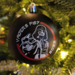 A black ornament hanging in a lit Christmas tree. On the bauble we see a stylized drawing of Darth Vader reaching out to us with the text "Vader's Fist 2022".