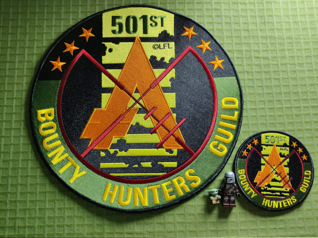 A large embroidered patch with the Bounty Hunters Guild logo. The colors is green and yellow (like Boba Fett). The symbol is a blaster crosshair aiming at the aurebesh symbol for galactic credits. The background is the famous killstripes from Boba Fett's helmet. There's six stars, symbolizing the six original bounty hunters from Empire Strikes Back. A regular sized patch and two lego figures is also visible, looking small besides the large patch.