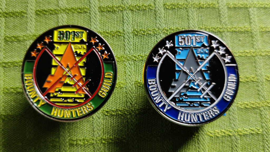 Two pins with the Bounty Hunters Guild logo. Only the colors are different. One is green and yellow (like Boba Fett), the other is blue and silver (like Jango Fett). The symbol is a blaster crosshair aiming at the aurebesh symbol for galactic credits. The background is the famous killstripes from Boba Fett's helmet. There's six stars, symbolizing the six original bounty hunters from Empire Strikes Back.