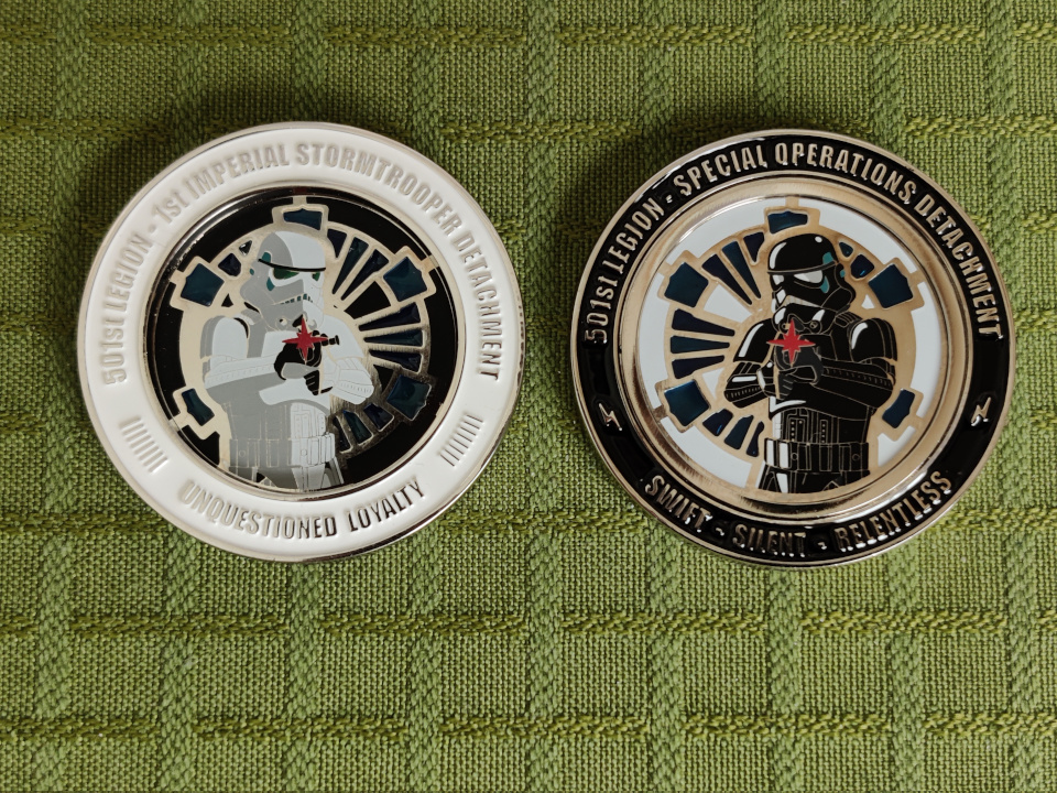 We see two coins. The coin to the left has the text "501st Legion. 1st Imperial Stormtrooper Detachment. Unquestioned Loyalty". The frame is white, the background is black and in the middle we see an imperial Stormtrooper in front of an imperial cog. The trooper is firing a blaster.The coin to the right has the text "501st Legion. Special Operations Detachment. Swift, Silent, Relentless." The frame is black, the background is white and in the middle we see an imperial Shadowtrooper in front of an imperial cog. The trooper is firing a blaster.The cog is blue, translucent glass. 