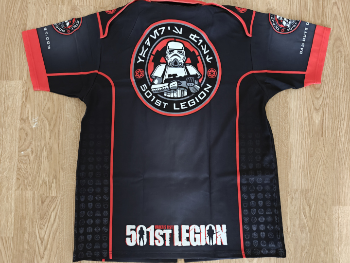 A dark club shirt, seen from the back. The shirt is laying flat on the floor.A large round 501st logo is printed on the top of the back, and a large 501st letter logo is printed on the bottom of the back.