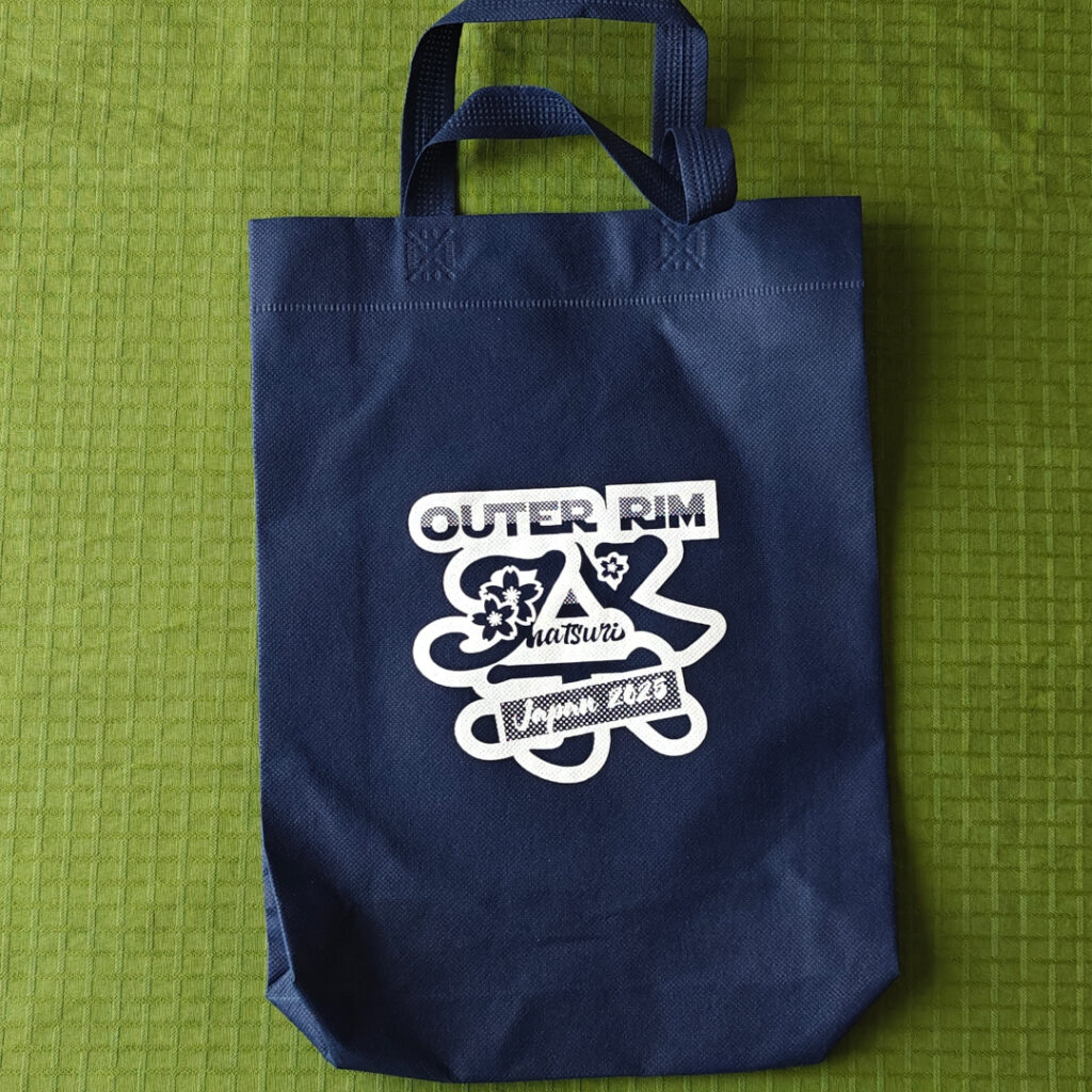 A blue bag with the bash logo in white. The logo has three lines. The top line: "Outer Rim". The middle line is a stylized japanese kanji with cherry blossoms, and also regular latin letters: "Matsuri". The bottom line is a box with letters: "Japan 2025".