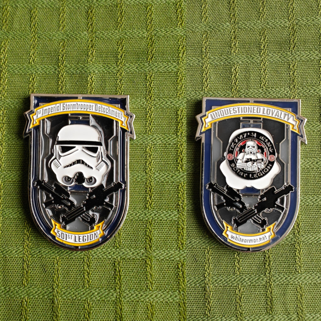 A coin shaped like a shield, seen from both sides. The front is the First Imperial Stormtrooper Detachment standard logo, with a stormtrooper helmet and two E11 blaster rifles in a cross. A banner above the helmet says "1st Imperial Stormtrooper Detachment", and a banner below the blaster rifles says "501st Legion". The back side has the 501st standard logo instead of the helmet. The top banner says "Unquestioned Loyalty" and the bottom banner says "whitearmor.net".